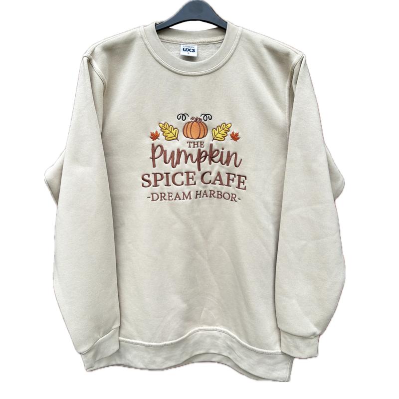 The Pumpkin Spice Café Official Embroidered Crew Neck Sweatshirt - A warm beige super-soft sweatshirt with embroidered details (a collegiate style centre slogan, a pumpkin & autumn leaves). Boxy fit, crew neckline, cuffed sleeves and bottom hem. 1